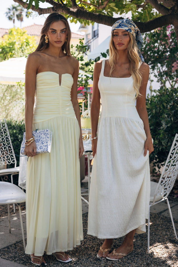 Two women in strapless dresses standing outdoors with greenery and chairs in the background.