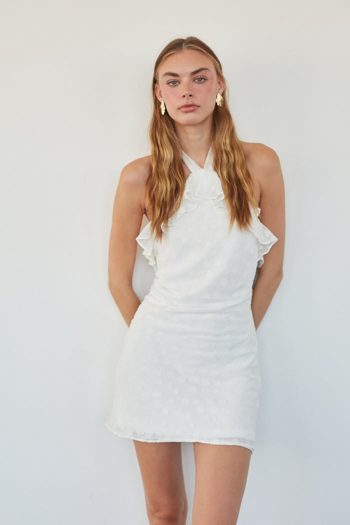 A woman modeling a white halter-neck mini dress with ruffle details and a textured fabric, set against a plain white background.
