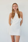 A woman modeling a white halter-neck mini dress with ruffle details and a textured fabric, set against a plain white background.