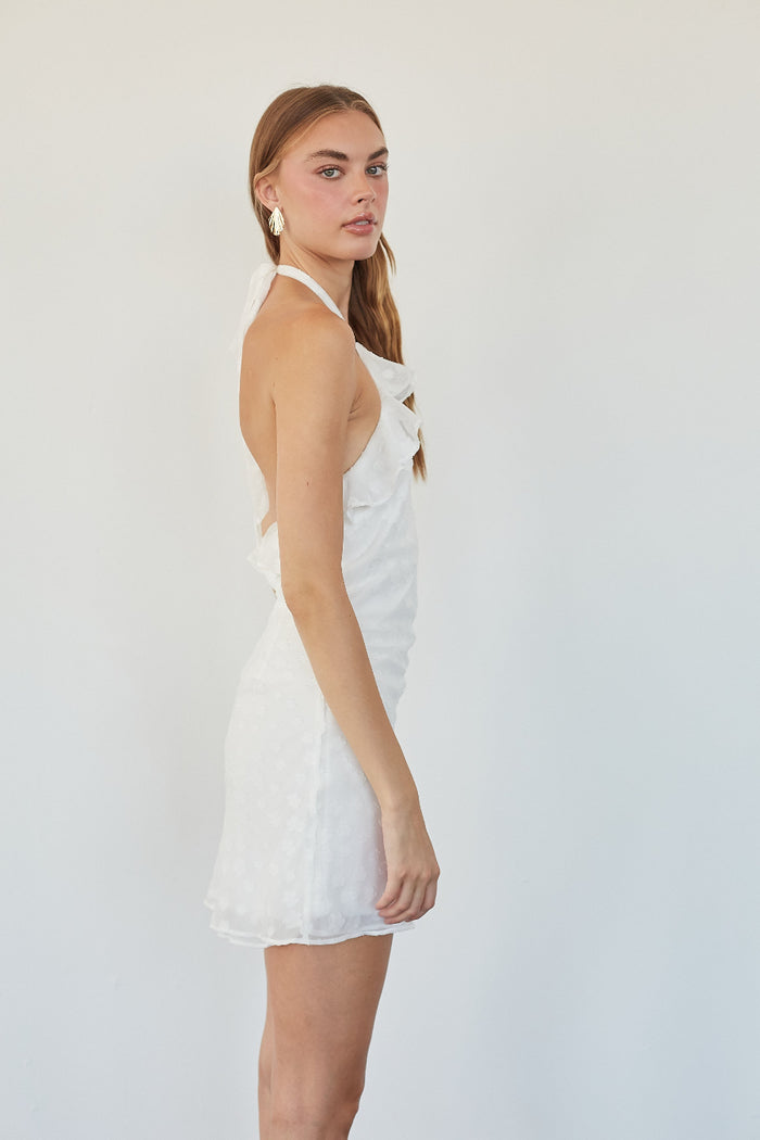 A woman modeling a white halter mini dress with a textured pattern, shown from a side-back angle against a plain white studio background.