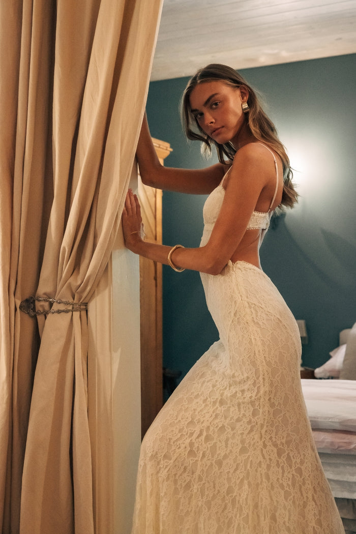 Woman in a white lace dress standing in a room with beige curtains.