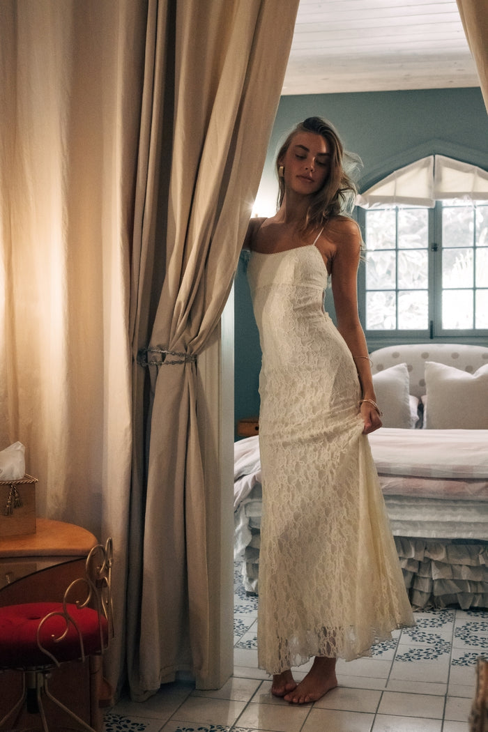 Woman in a white lace dress standing behind a curtain in a room with a couch and window.