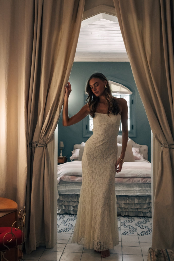 Woman in a white dress standing in a bedroom with curtains drawn back.