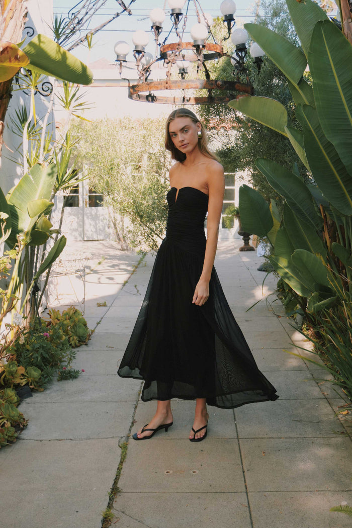 Woman in a black strapless dress standing in a garden setting with plants and a chandelier.