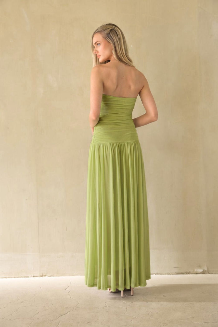 A woman stands with her back to the camera wearing a strapless lime green maxi dress with a ruched bodice and flowing pleated skirt, set against a neutral beige textured wall.