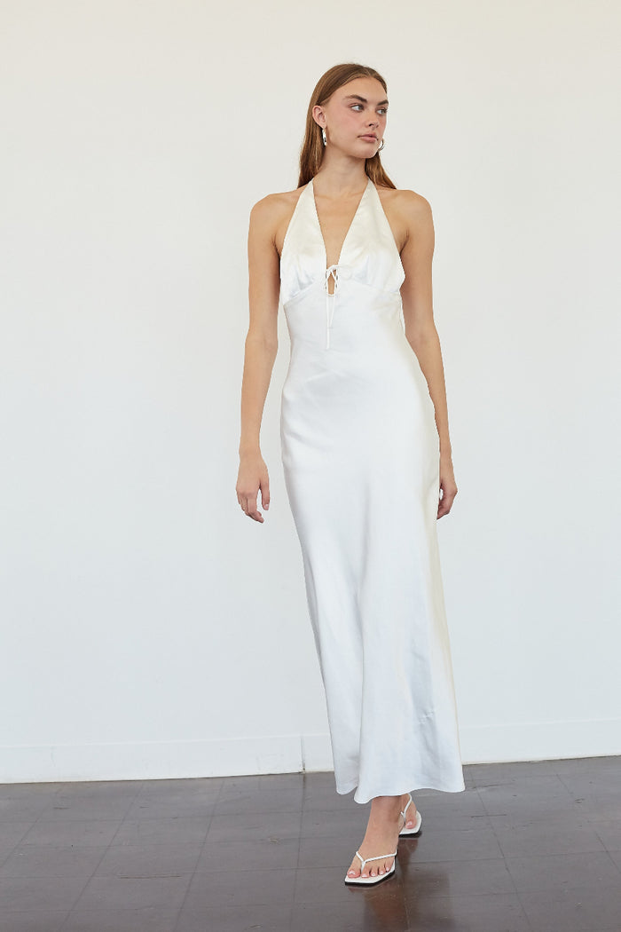 A woman modeling a white halter-neck maxi dress with a deep plunging V-neckline and tie detail at the bust, set against a plain white wall and dark gray floor.