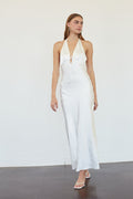 A woman modeling a white halter-neck maxi dress with a deep plunging V-neckline and tie detail at the bust, set against a plain white wall and dark gray floor.