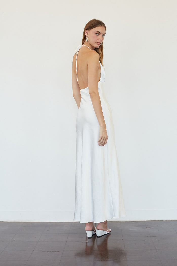 A woman modeling a white satin maxi dress with a deep back and thin straps, standing against a plain white wall on a dark wooden floor.