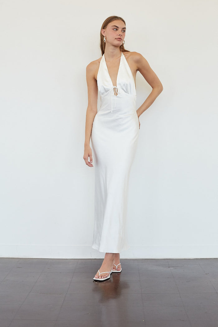 A woman modeling a white halter-neck maxi dress with a plunging neckline and tie-front detail, standing against a plain white wall on a dark floor.