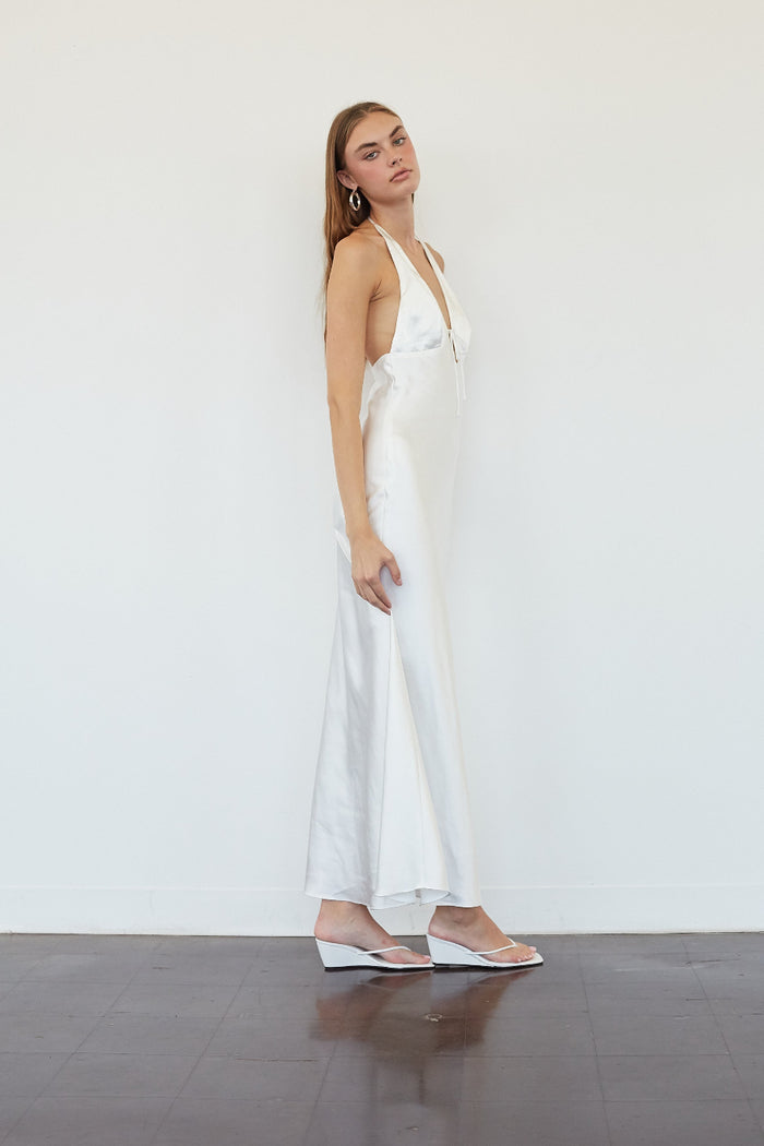 A woman modeling a white satin halter maxi dress with a side slit, paired with white wedge sandals, standing against a plain white wall.