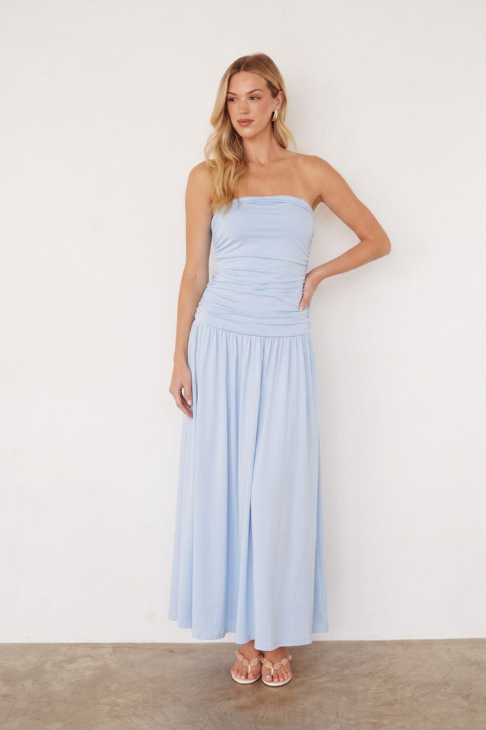 blue-image | A woman modeling a light blue strapless maxi dress with a ruched bodice and flowing skirt, standing against a plain off-white wall.