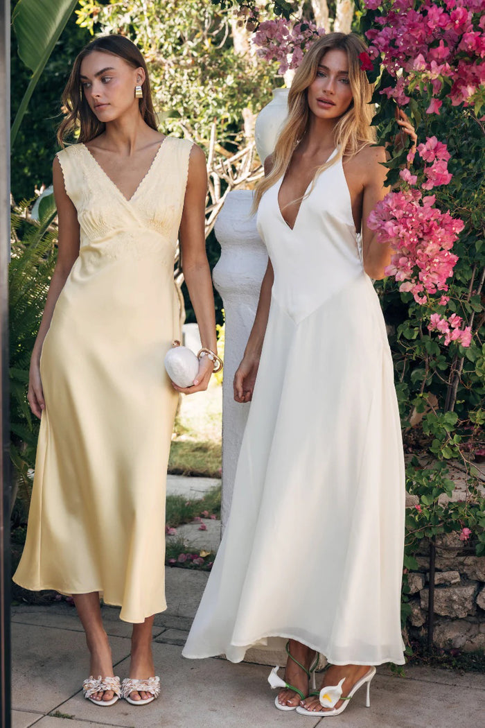 Two women in white dresses standing outdoors with greenery and pink flowers in the background.