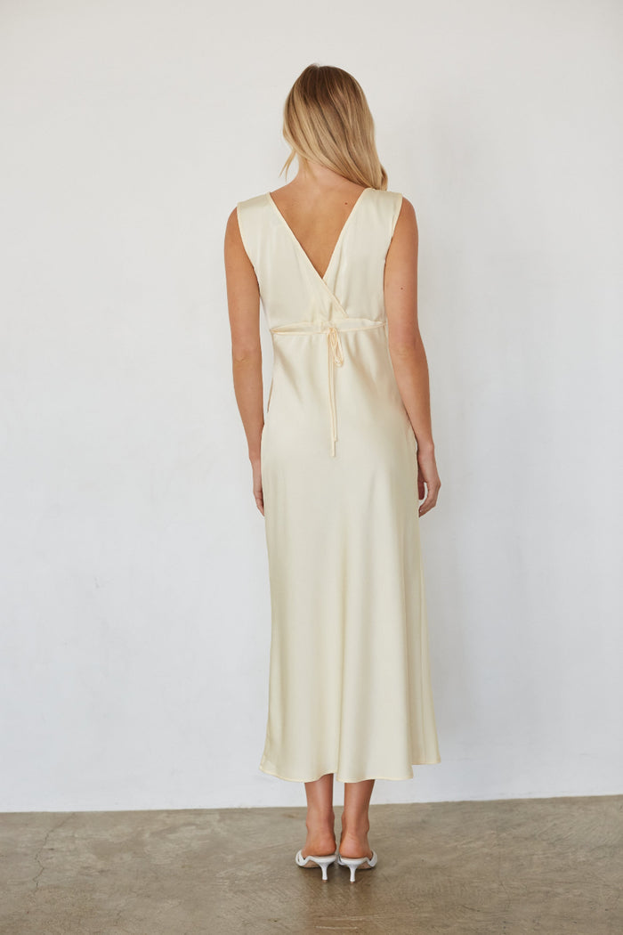 A woman viewed from the back wearing a sleeveless cream maxi dress with a V-neckline and a tie detail at the waist, paired with white high heels against a plain white wall.