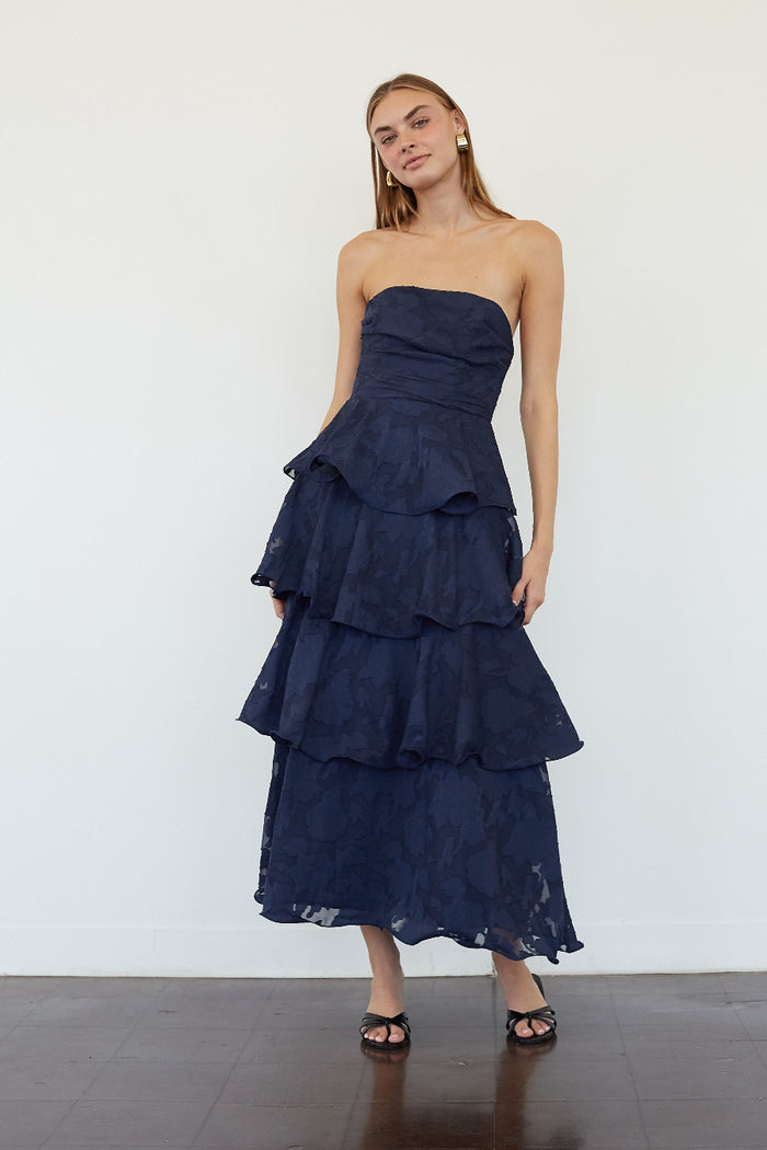 blue-image | A woman modeling a strapless navy blue maxi dress with a ruched bodice and a tiered ruffle skirt, standing against a plain white wall.