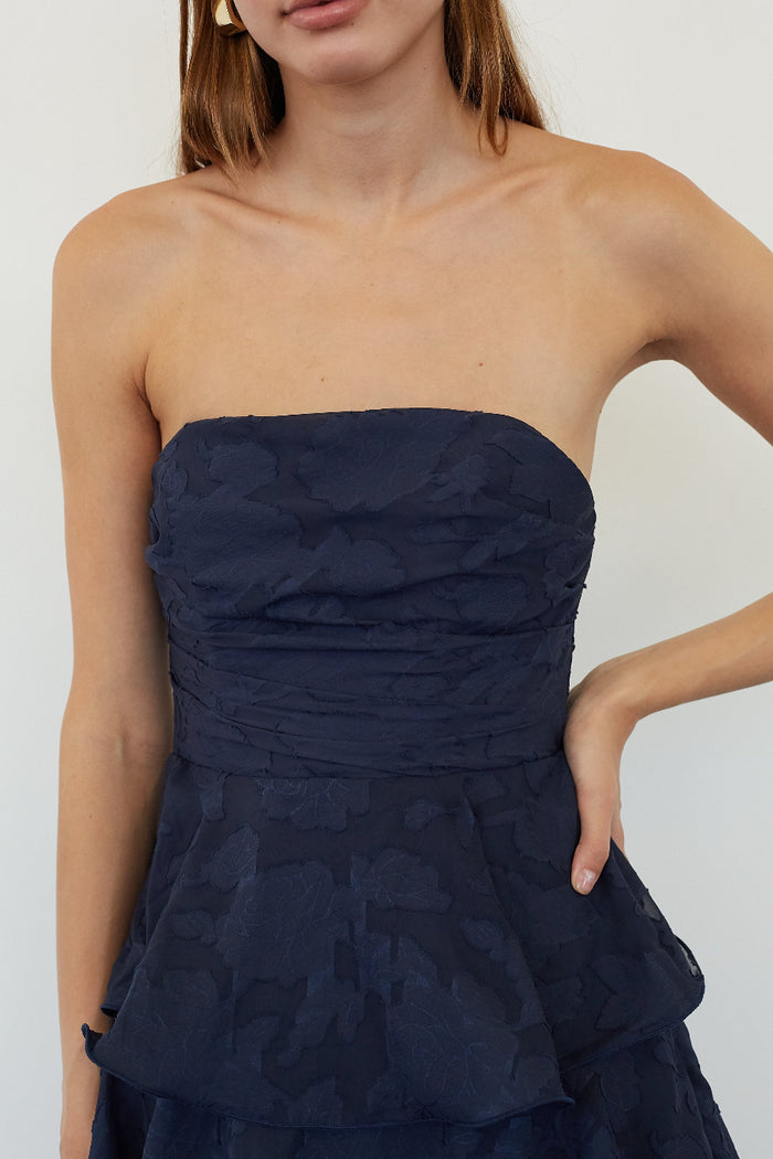A close-up of a model wearing a strapless navy blue dress with a textured, tonal floral jacquard pattern and a peplum waist.
