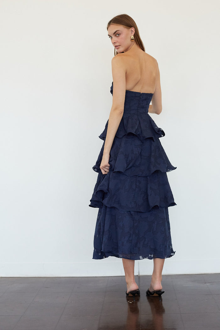 A woman modeling a strapless navy blue midi dress with a structured waist and layered ruffle skirt, shown from the back against a plain white wall.