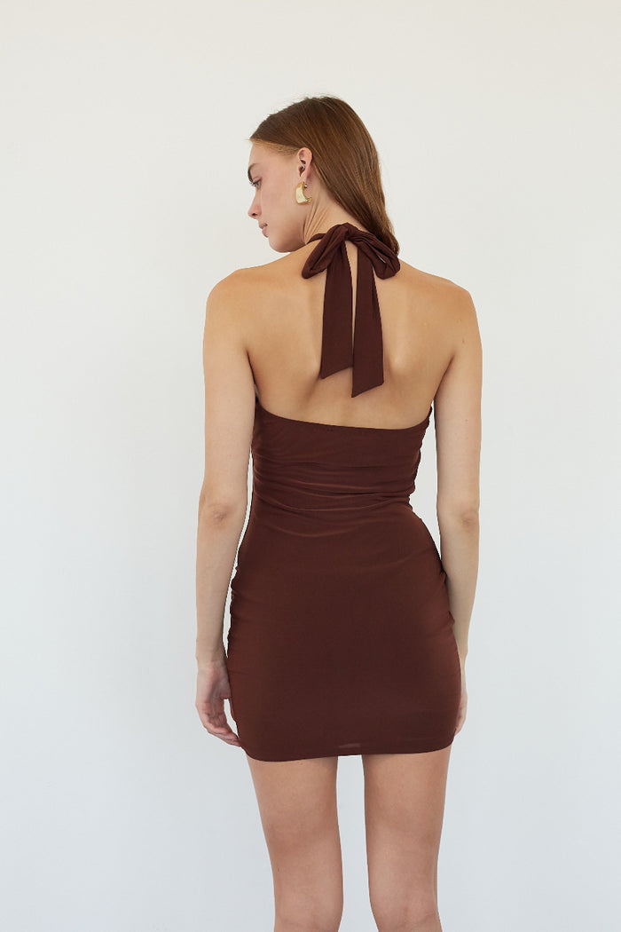 A woman shown from the back wearing a dark brown halter mini dress with a ruched bodice and side cutouts, against a plain white studio background.