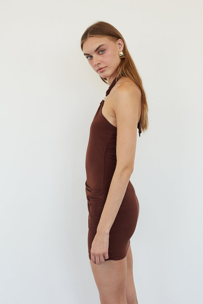 A woman modeling a sleeveless, halter-style brown mini dress against a plain white studio background.