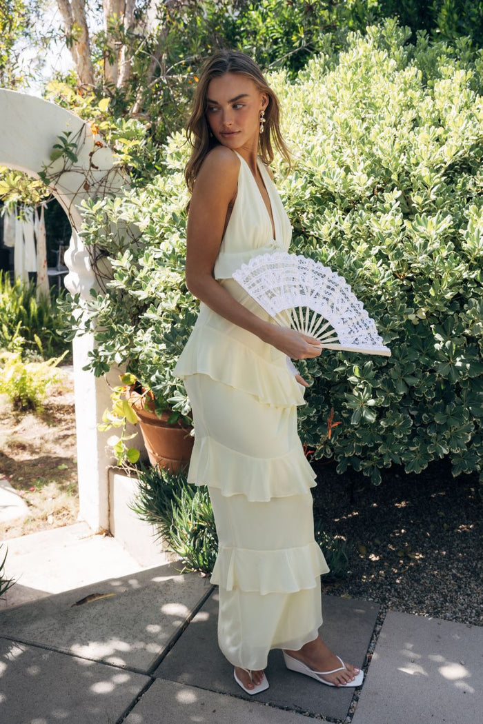 Woman in a light yellow dress holding a white fan outdoors.