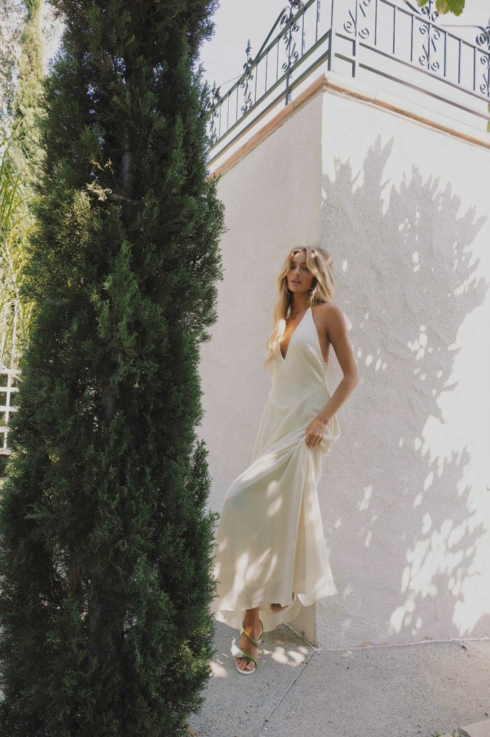 Woman in a ivory dress standing next to a tall green tree with a white wall and black railing in the background.