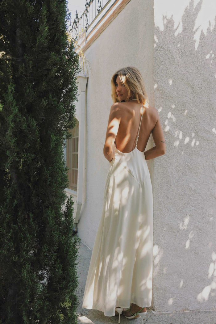 Woman in a ivory dress standing against a white wall with greenery around.