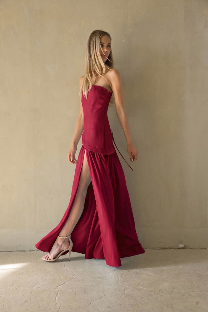 A woman modeling a strapless, floor-length magenta dress with a high thigh slit, standing against a textured beige wall.
