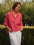 Woman wearing a red shirt and white pants standing outdoors with greenery in the background