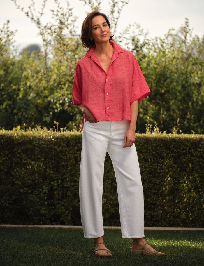 Woman wearing a red shirt and white pants standing outdoors with greenery in the background