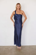 blue-image | A woman modeling a navy blue satin one-shoulder maxi dress against a plain white wall and concrete floor.