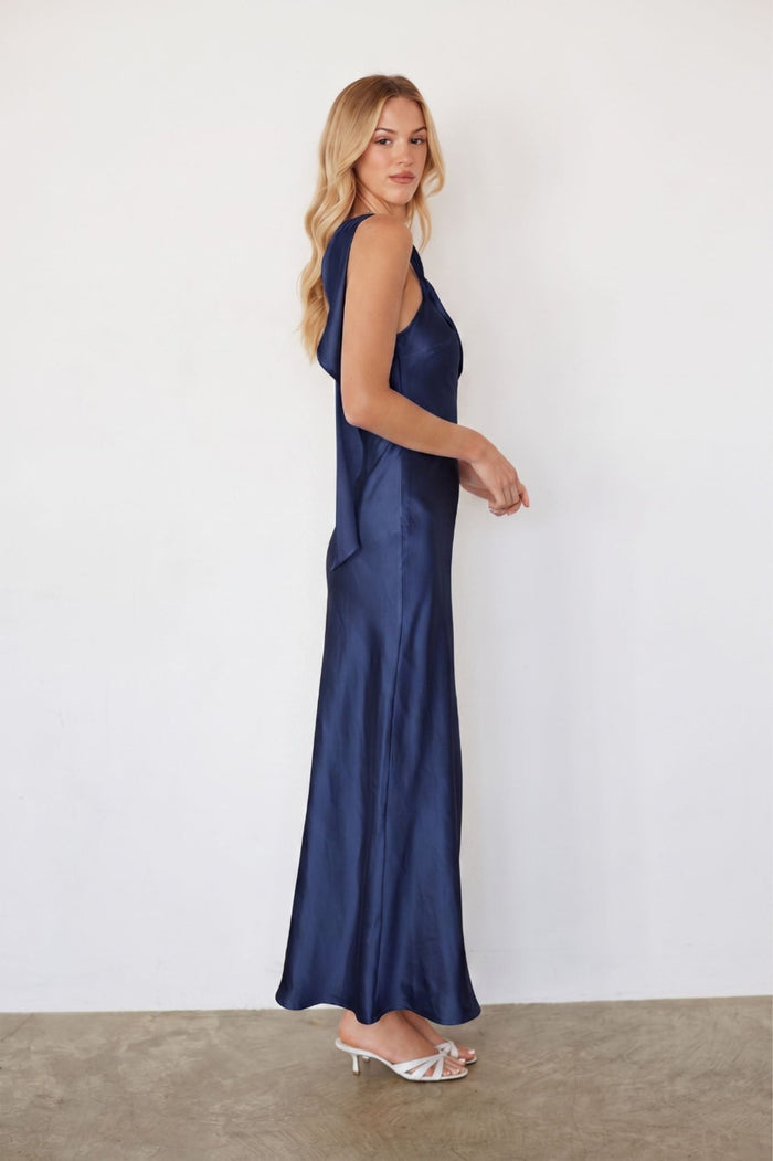 A woman modeling a sleeveless navy blue satin maxi dress with a deep V-neckline, shown from a side-back angle against a plain white wall and concrete floor.