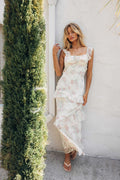 Woman in a floral dress standing against a white wall with greenery.