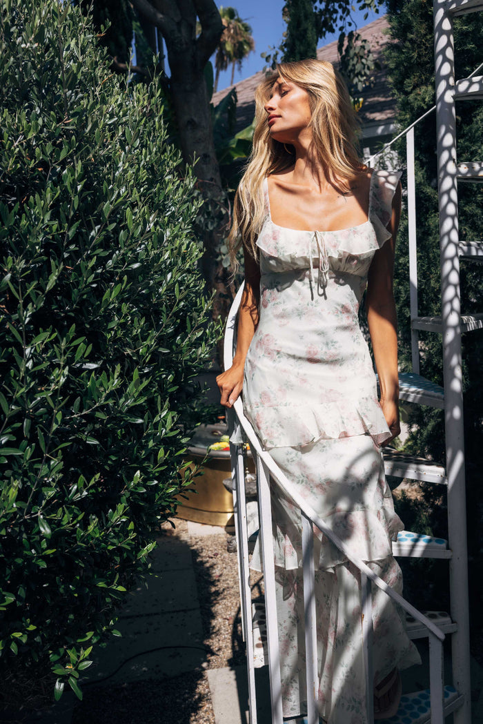 Woman in a floral dress standing outdoors with greenery and a ladder in the background
