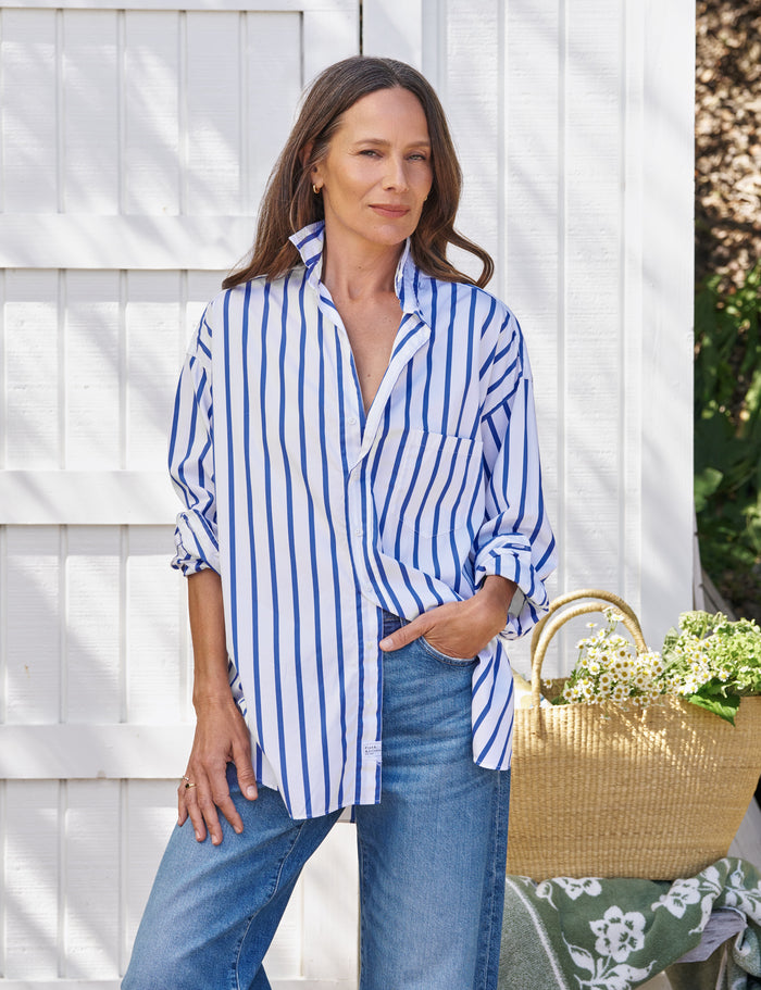 Woman wearing a blue and white striped shirt and jeans, standing next to a woven basket with flowers against a white wooden fence.