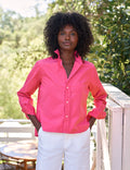 Woman wearing a bright pink shirt and white pants standing outdoors with greenery in the background