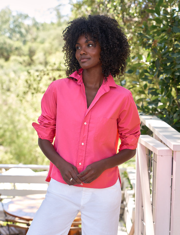 Woman wearing a bright pink shirt and white pants standing outdoors with greenery in the background