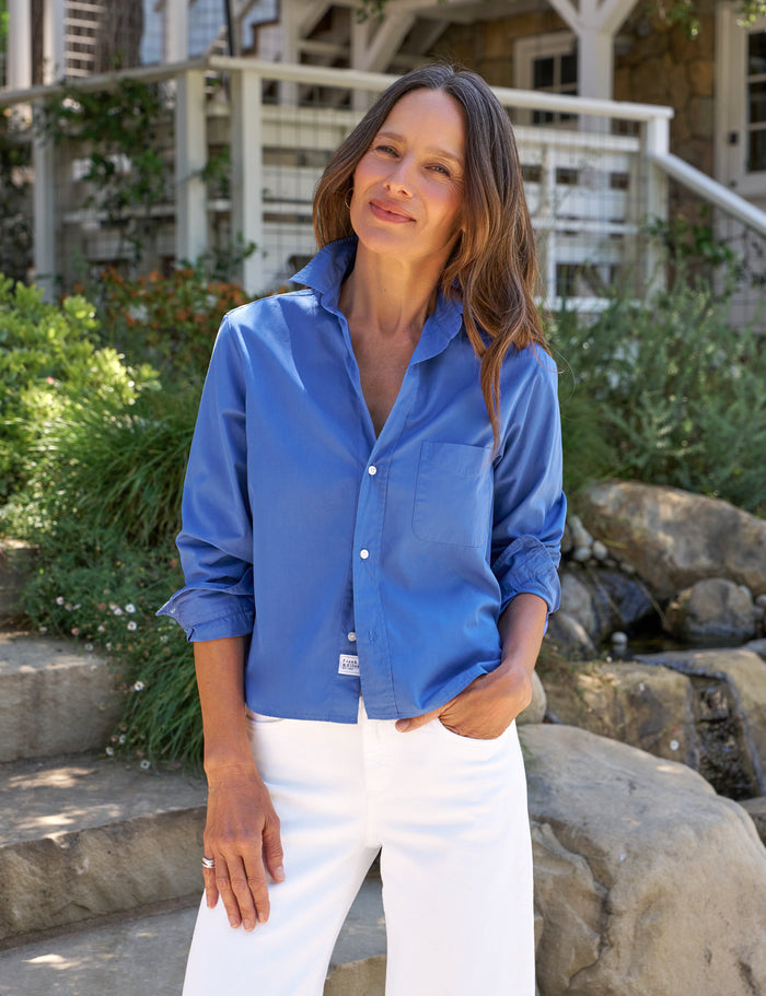 Woman wearing a blue shirt and white pants standing outdoors with greenery and a building in the background