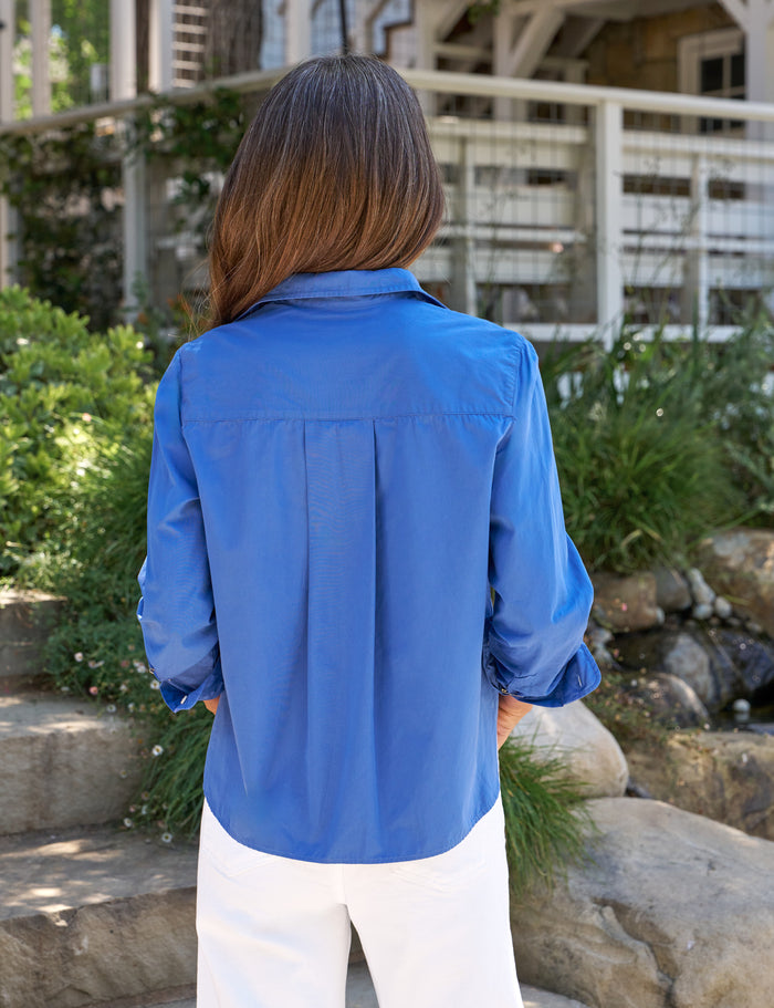 Person wearing a blue shirt standing outdoors with greenery and a building in the background