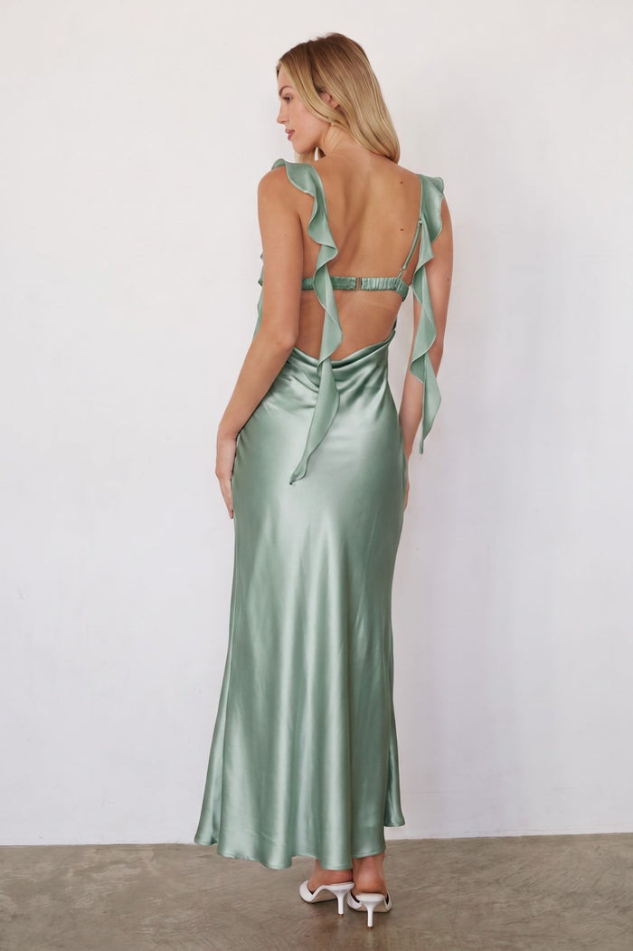 A woman stands with her back to the camera wearing a satin-like sage green maxi dress with a ruffled open back and tie details, paired with white heeled sandals against a plain light wall.