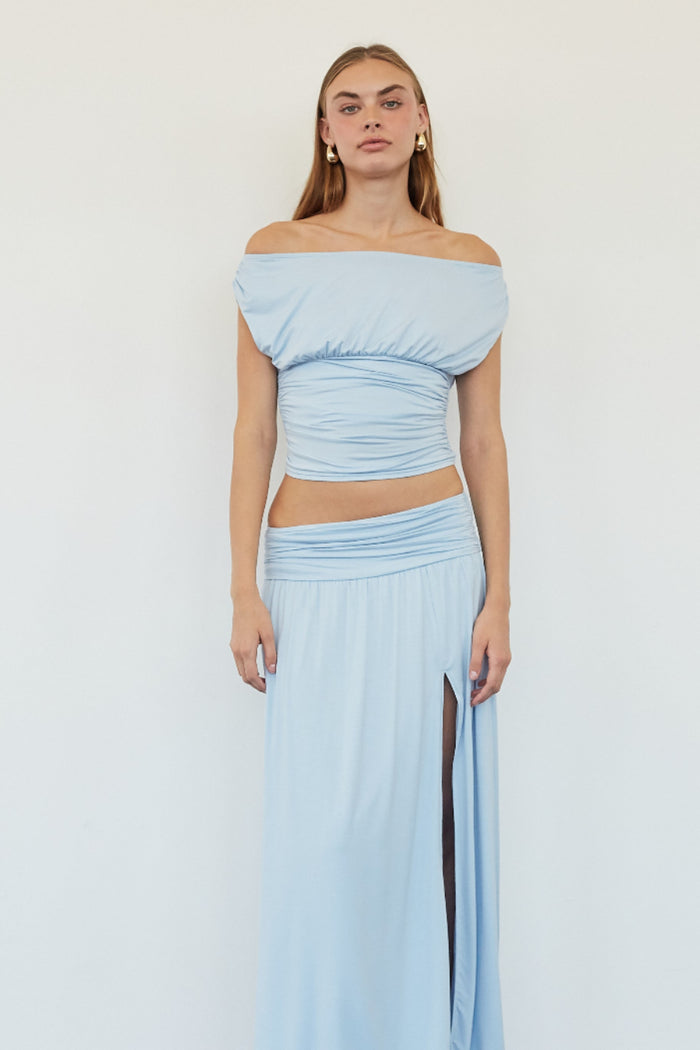 A woman modeling a light blue two-piece outfit consisting of an off-the-shoulder cropped top and a matching high-slit maxi skirt, standing against a plain white wall.