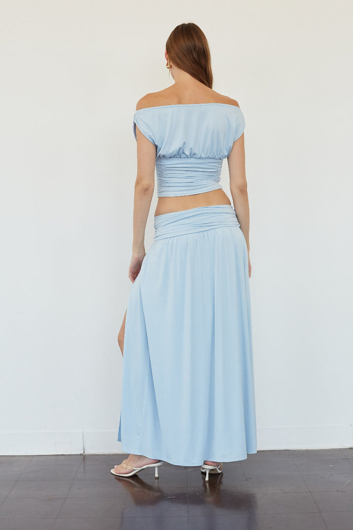 A woman stands with her back to the camera wearing a light blue off-the-shoulder two-piece outfit with a cropped top and a long skirt, paired with clear high-heeled sandals, against a plain white wall.