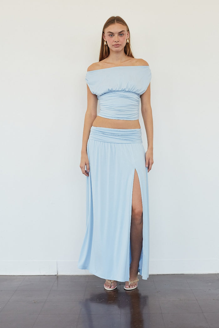 A woman modeling a light blue two-piece outfit consisting of an off-the-shoulder crop top and a matching high-slit maxi skirt, standing against a plain white wall.