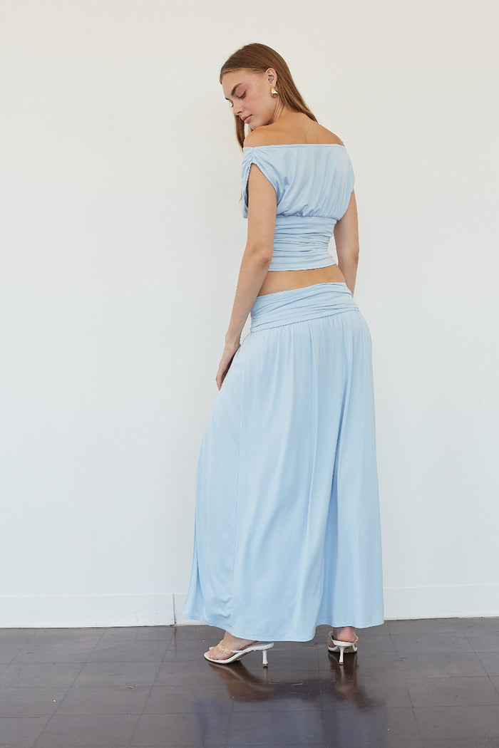 A woman modeling a light blue two-piece outfit consisting of an off-the-shoulder crop top and matching wide-leg maxi skirt, standing against a plain white wall.
