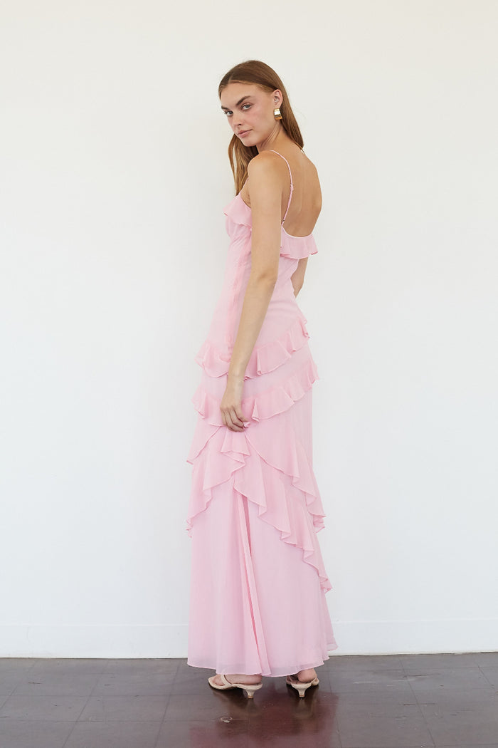 A woman modeling a light pink ruffled maxi dress with spaghetti straps and an open back, standing against a plain white wall.
