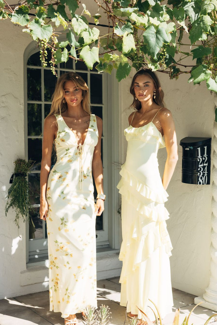 yellow-image | Two women in elegant dresses standing under a tree with a white building in the background.