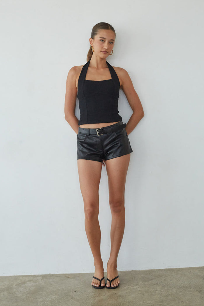 Woman wearing a black halter top and leather shorts against a white wall.