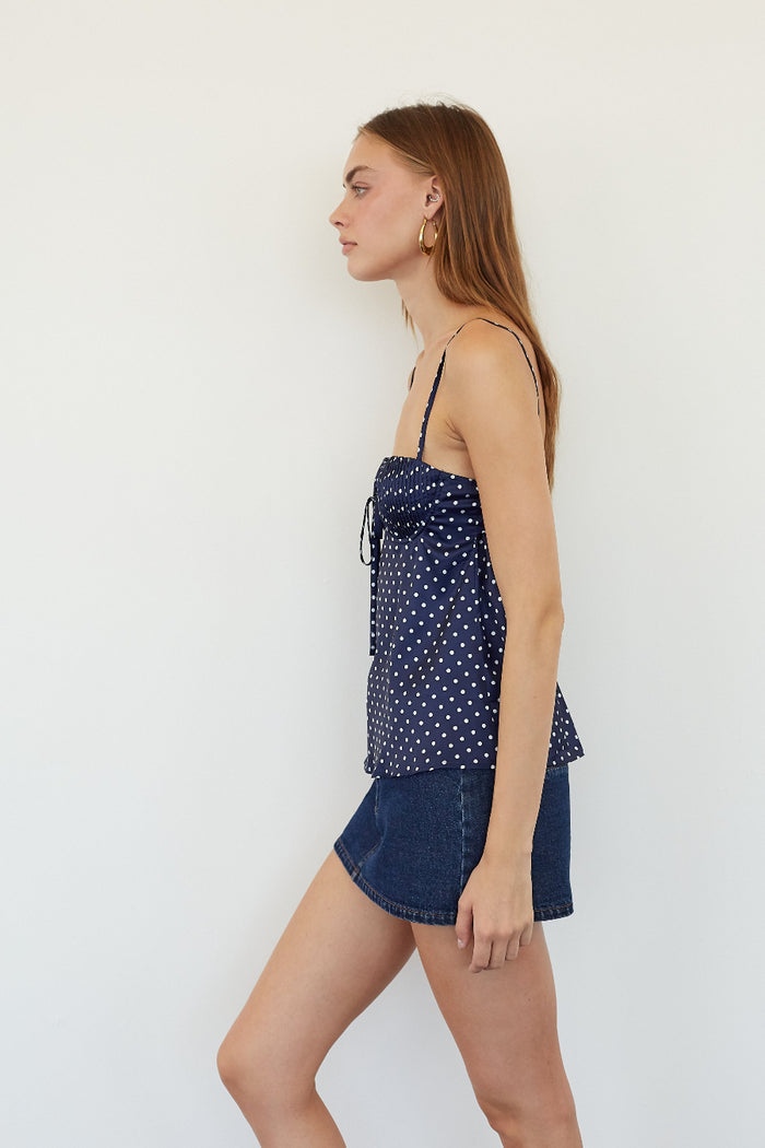 A woman in side profile wearing a navy blue camisole top with white polka dots and dark denim shorts, standing against a plain white wall.