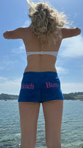 A woman stands with her back to the camera outdoors by the water, wearing blue swim shorts with pink text reading 'Beach Bum' and a white bikini top. The background shows blue sky and water.