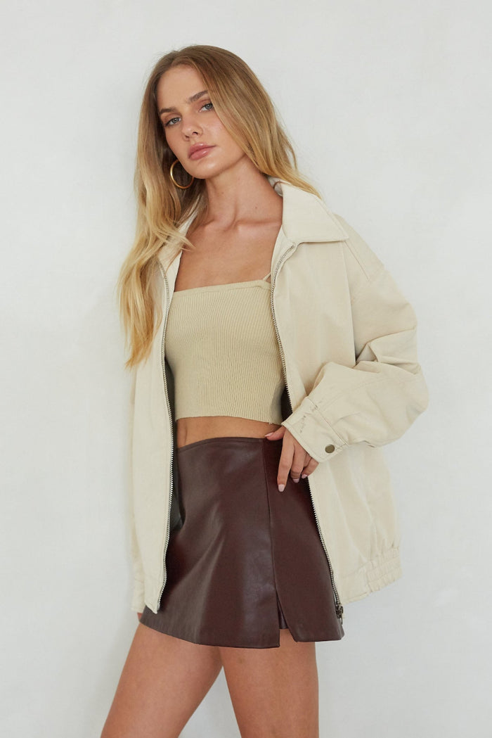 Woman wearing a beige jacket, beige top, and brown leather skirt against a white background