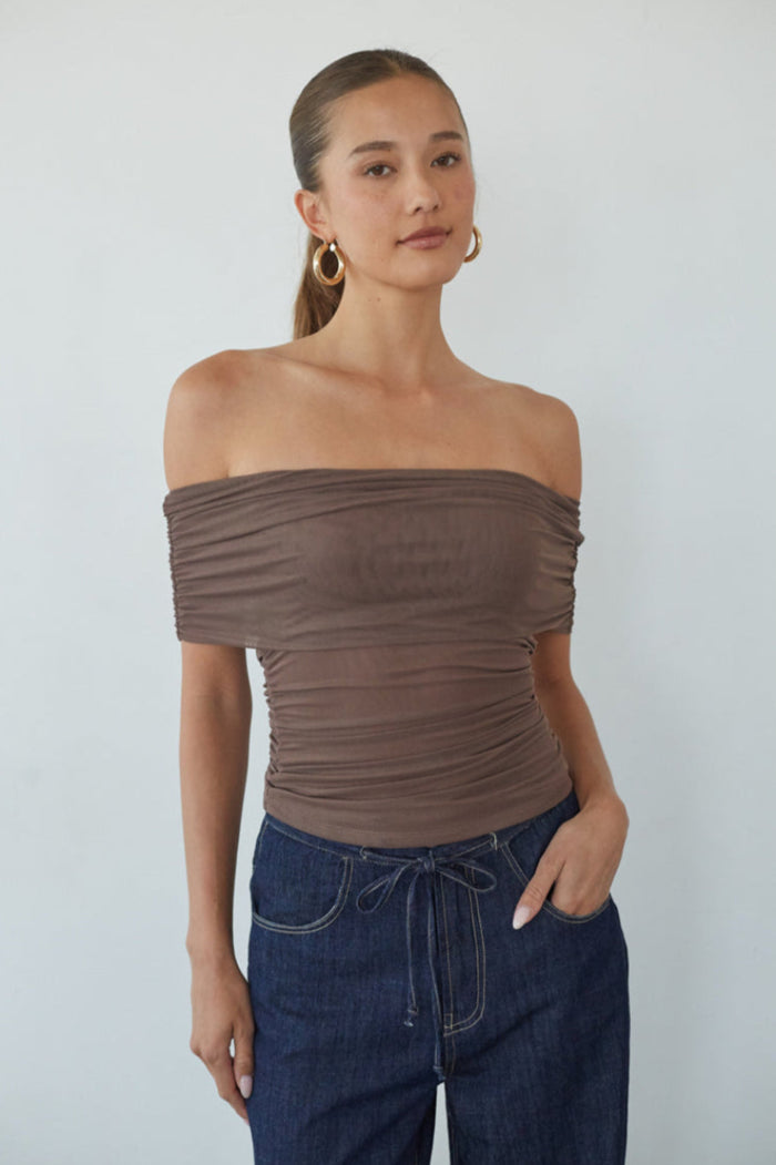 brown-image | Woman wearing a brown off-shoulder top and blue jeans against a plain background