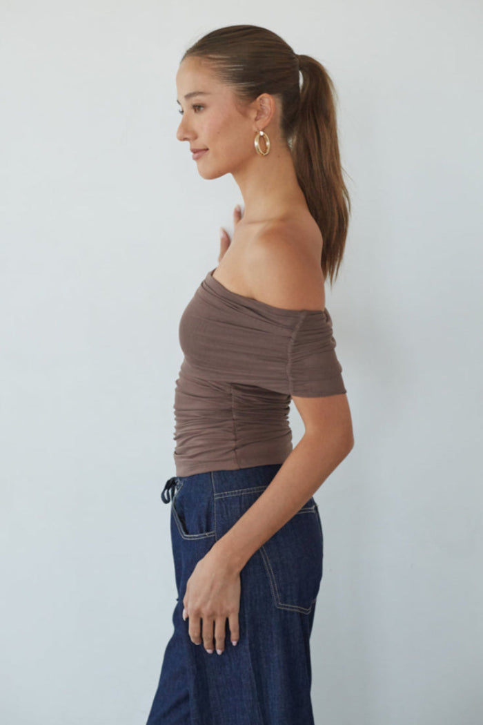 Woman wearing a brown off-shoulder top and blue pants against a white background